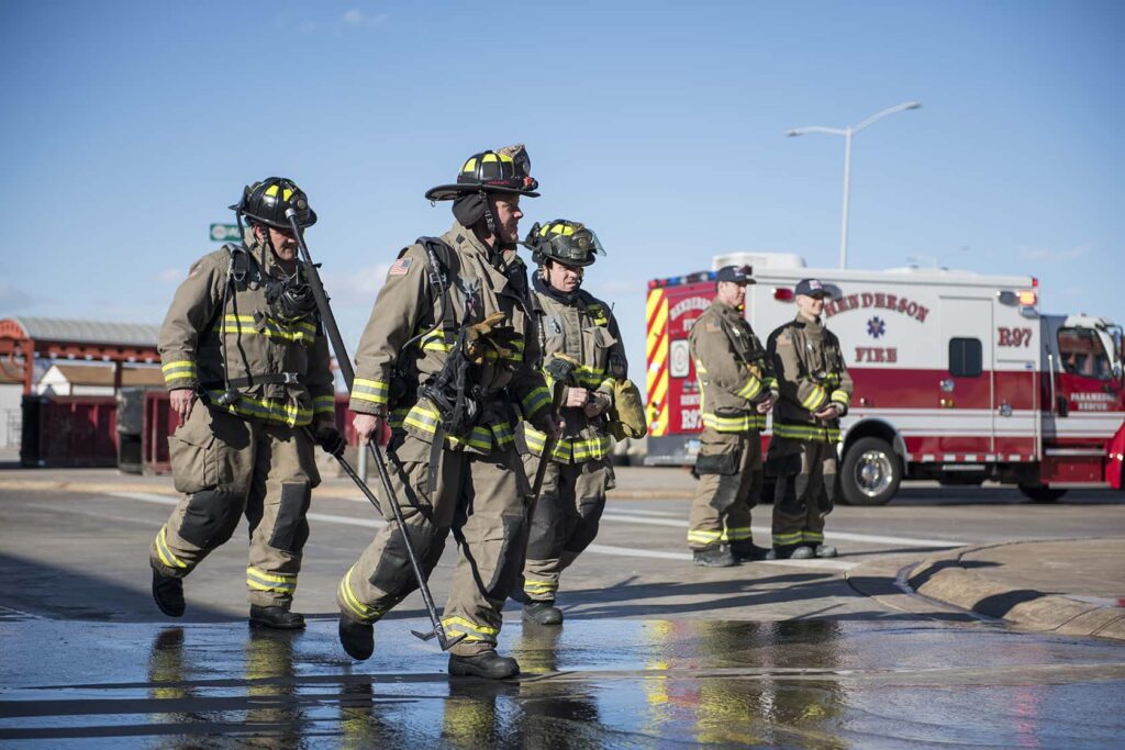 Four firefighters in full gear walk across a wet street, carrying tools. A fire truck is parked nearby, and the sky is clear.