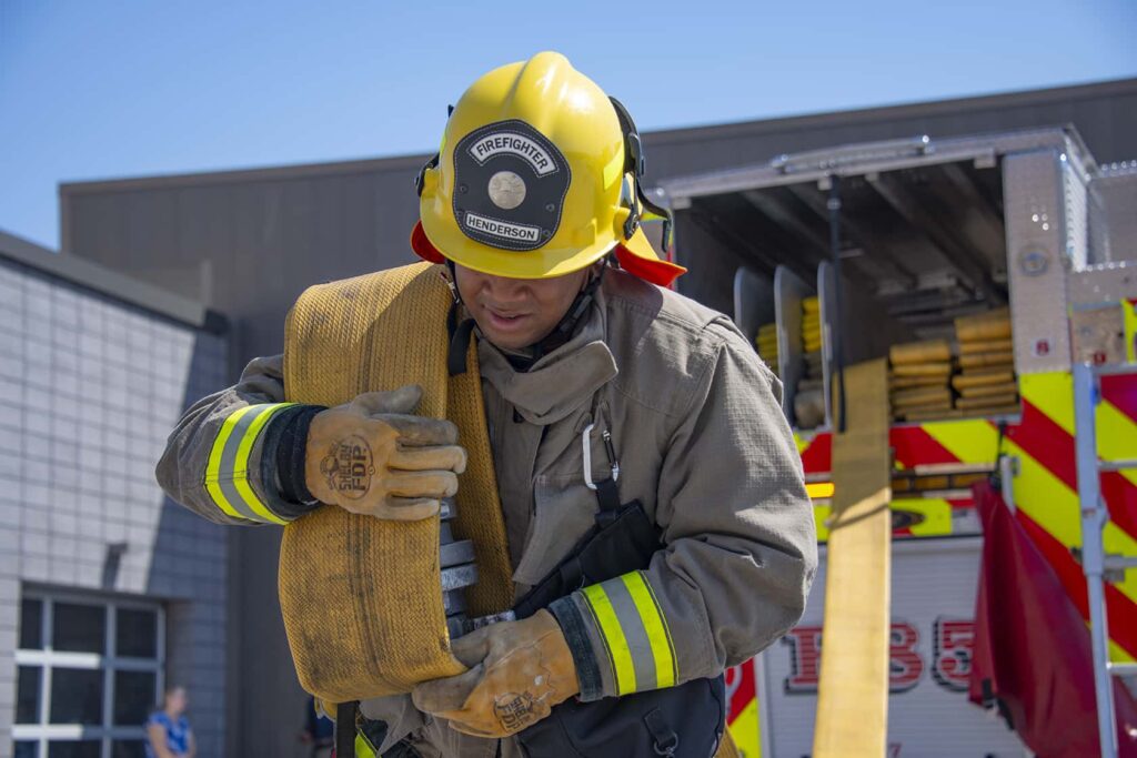 A firefighter wearing protective gear and a yellow helmet carries a rolled fire hose in front of a fire truck outside a fire station.