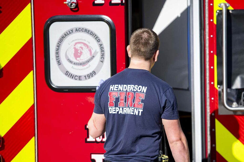 A person wearing a “Henderson Fire Department” shirt stands near the open door of a red emergency vehicle with yellow stripes and an accreditation seal on the door.