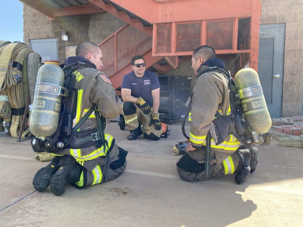 Two firefighters in full gear kneel on the ground, listening to an instructor in a navy shirt and sunglasses outside under a metal staircase, with equipment and hoses nearby.