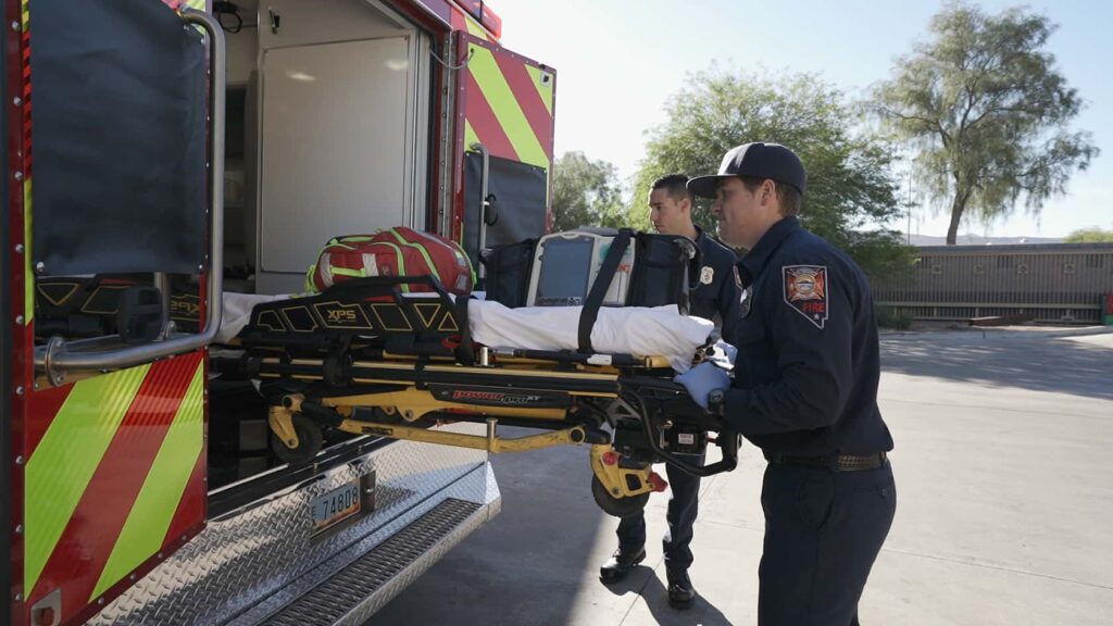 Two paramedics in uniform load an empty stretcher into the back of an ambulance on a sunny day, with trees and a building visible in the background.