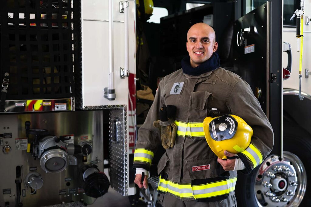 A firefighter in full gear smiles while holding a yellow helmet, standing next to a fire truck equipped with hoses and equipment.