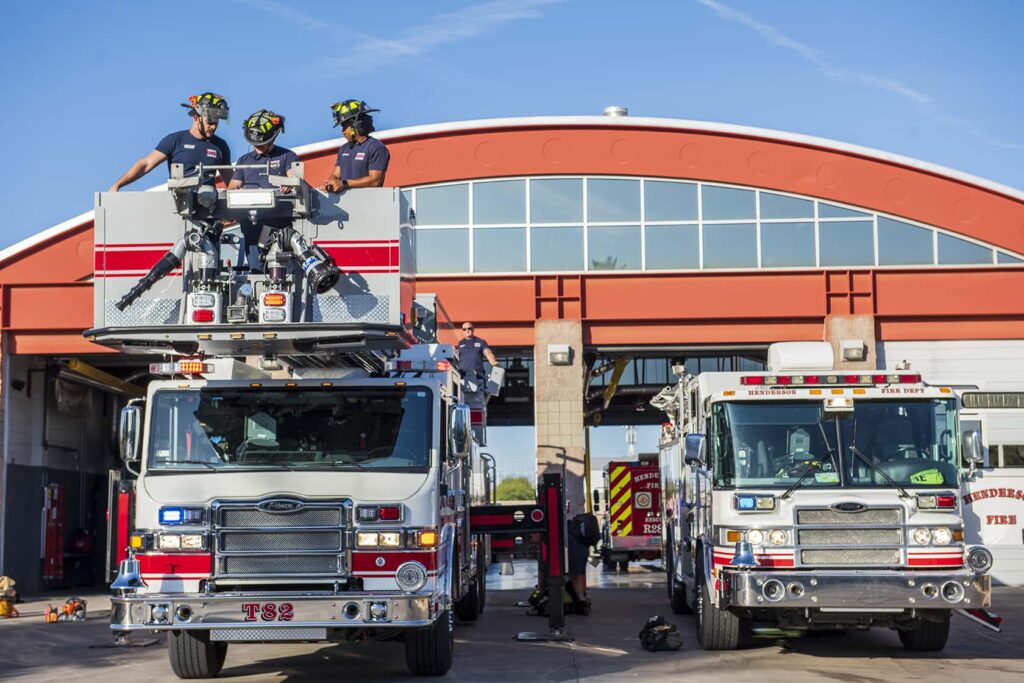 Two fire trucks are parked outside a fire station. Three firefighters stand on top of one truck’s extended platform, while others stand nearby. The fire station has a large arched roof and garage doors.