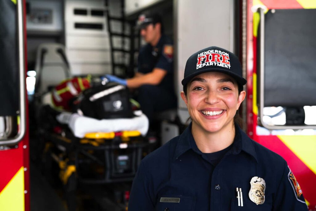 A smiling firefighter in uniform and a Henderson Fire Department hat stands in front of an ambulance, with another firefighter tending to a stretcher inside the vehicle.
