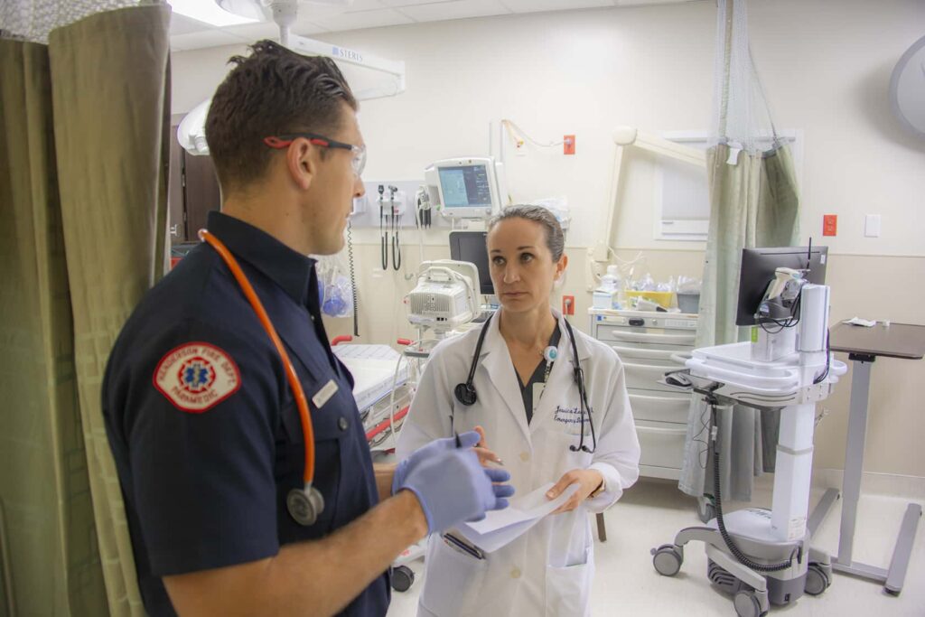 A paramedic and a doctor stand in a hospital room, engaged in conversation. The doctor holds a clipboard, and medical equipment surrounds them. Both wear stethoscopes around their necks.