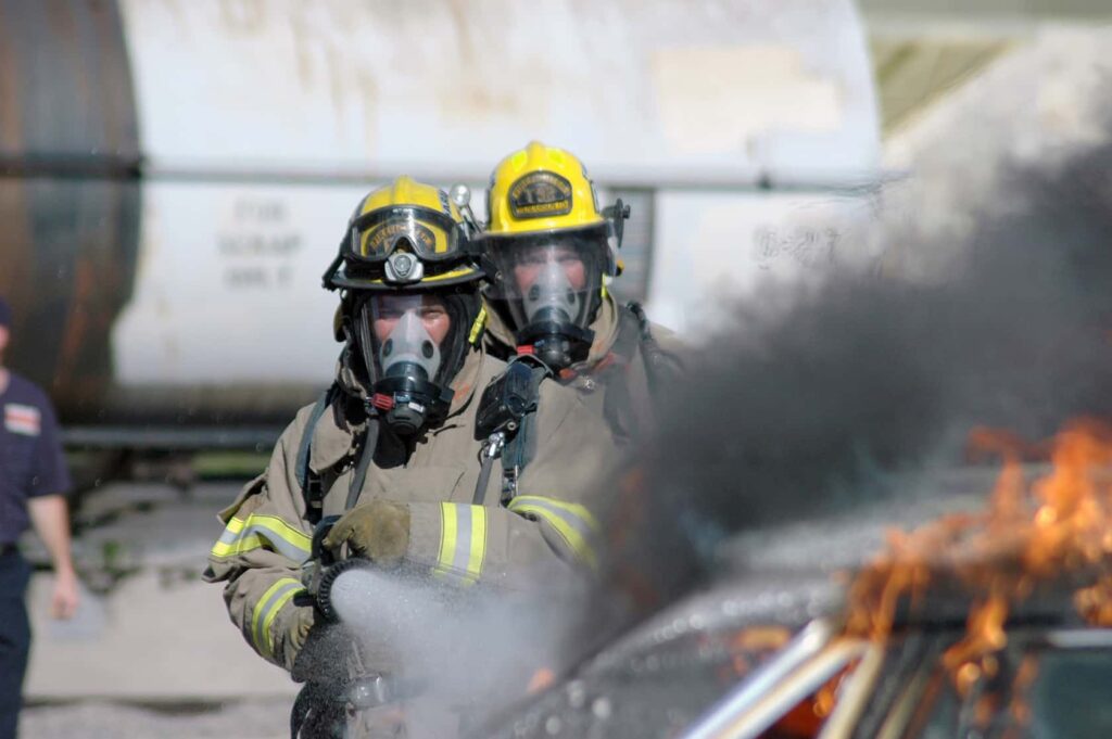 Two firefighters in full gear and helmets use a hose to extinguish a burning car, with thick black smoke rising. A large tank and another person are visible in the background.