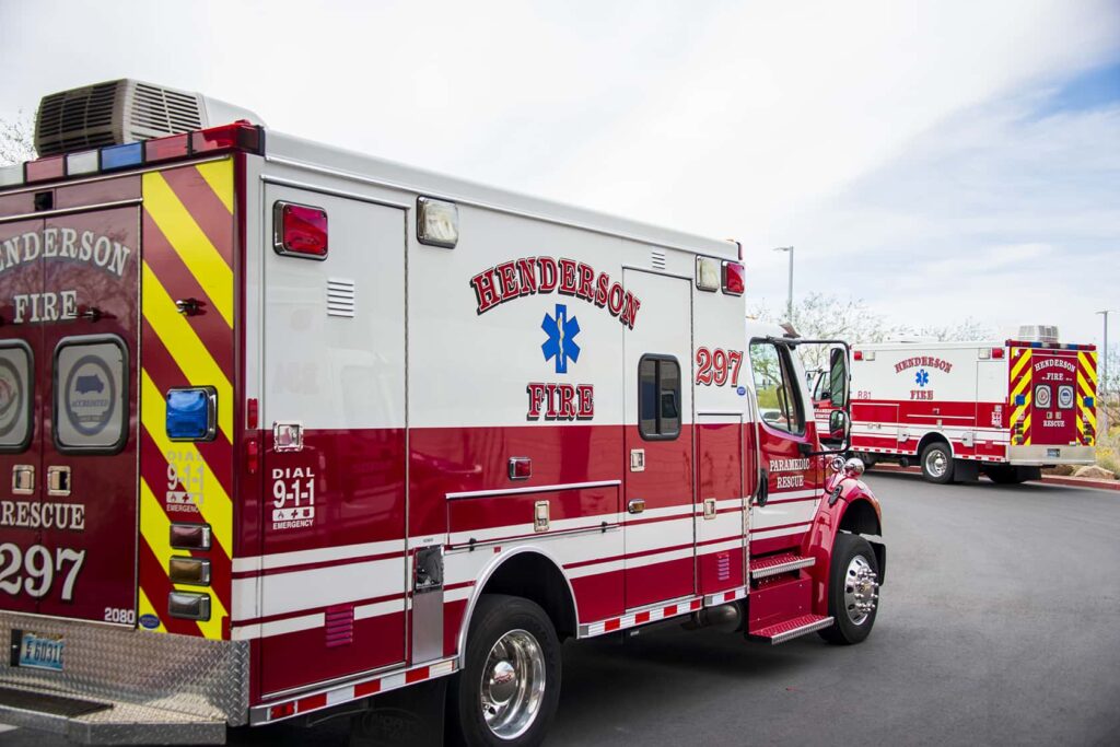 Two red and white Henderson Fire Department ambulances are parked outdoors on a paved surface under a partly cloudy sky. The vehicles have emergency symbols and stripes, with one in the foreground and one in the background.