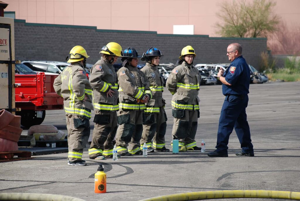 A group of five firefighters in full gear stand in a line outdoors, listening to an instructor in a navy uniform. They are in a parking lot with firefighting equipment nearby.