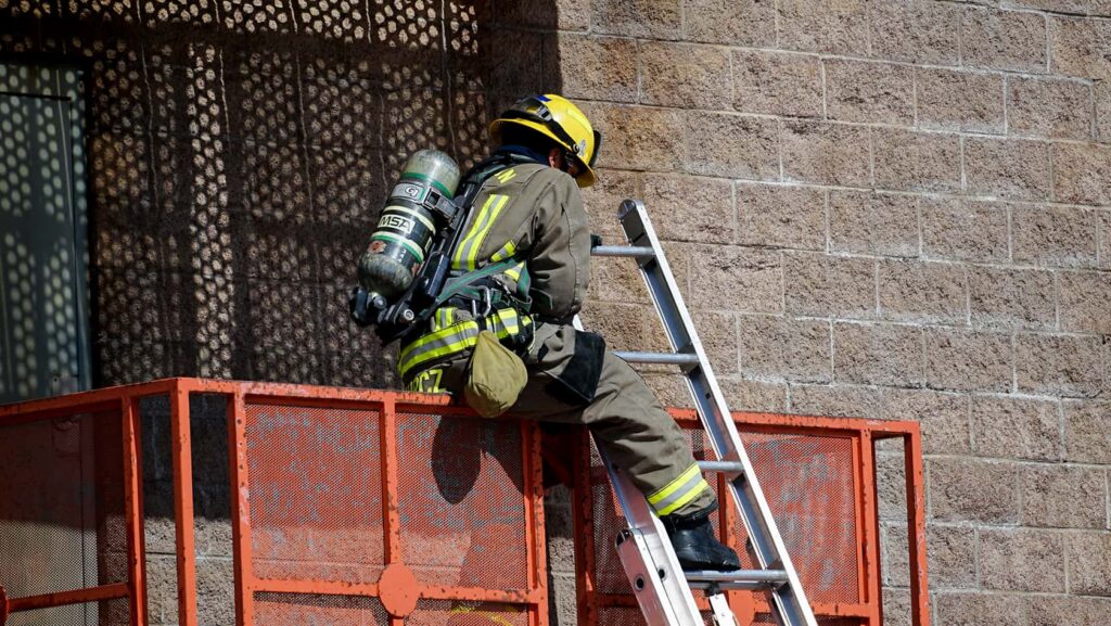 A firefighter in full gear and helmet climbs a metal ladder onto a raised platform beside a brick building, with sunlight casting shadows on the wall.