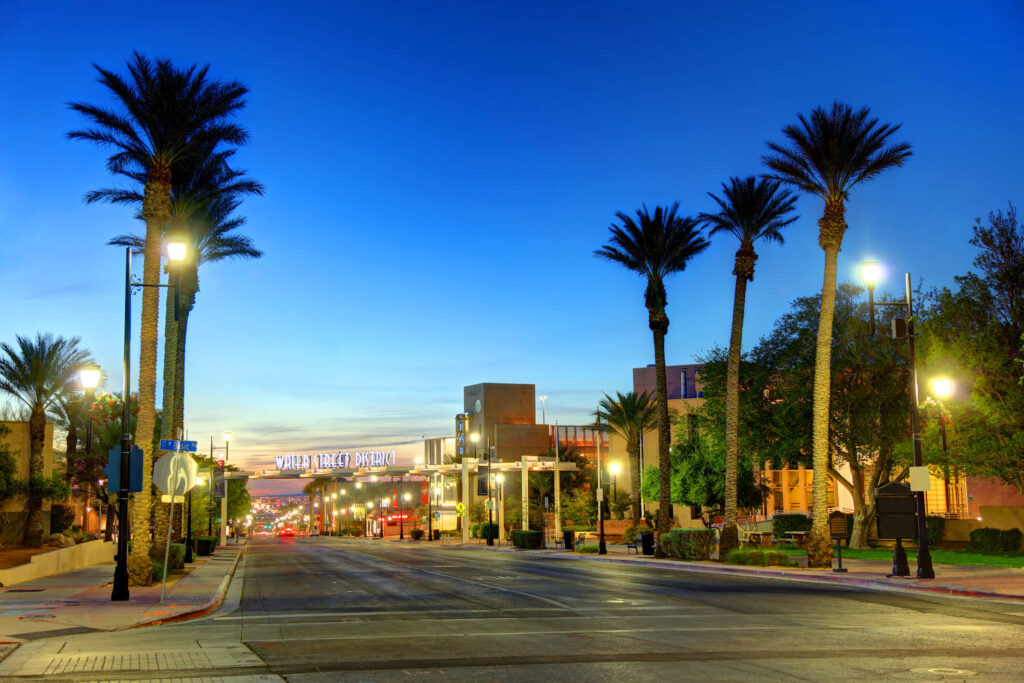 A wide, clean street lined with tall palm trees and streetlights at dusk, leading toward the “Water Street District” sign in a calm urban area with a clear, twilight sky.