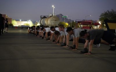 A group of people outdoors at dusk perform plank exercises in a row on pavement, with industrial equipment and lights in the background.