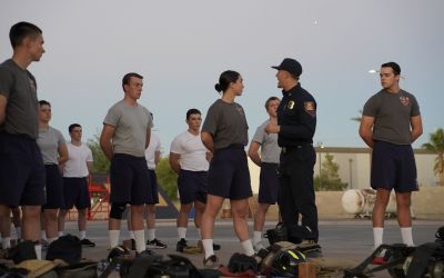 A group of trainees in athletic uniforms stand in formation outdoors while a uniformed instructor speaks to them. Firefighting gear is laid out on the ground in front of the group.