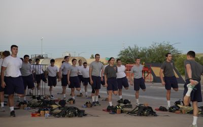 A group of young people in athletic wear stretch outdoors in formation, each holding one foot behind them. Equipment and bags are scattered on the ground. The sky is clear, and some trees and structures are visible in the background.